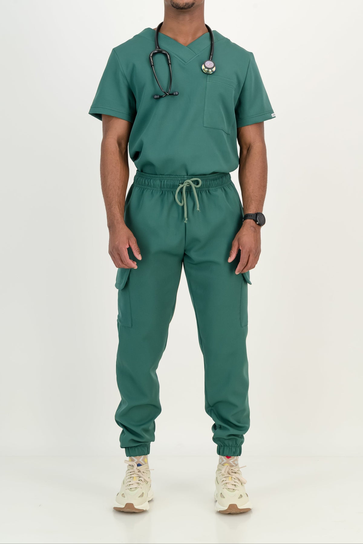 A man wearing a matching green medical scrub set with a V-neck top, cargo pants, and a stethoscope, standing against a plain white studio background.