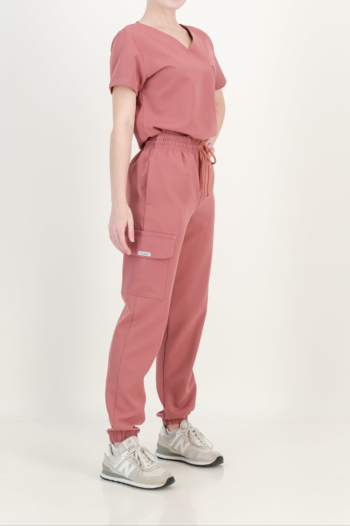 A woman modeling a matching dusty rose scrub top and jogger pants set, paired with light gray sneakers, against a plain off-white studio background.