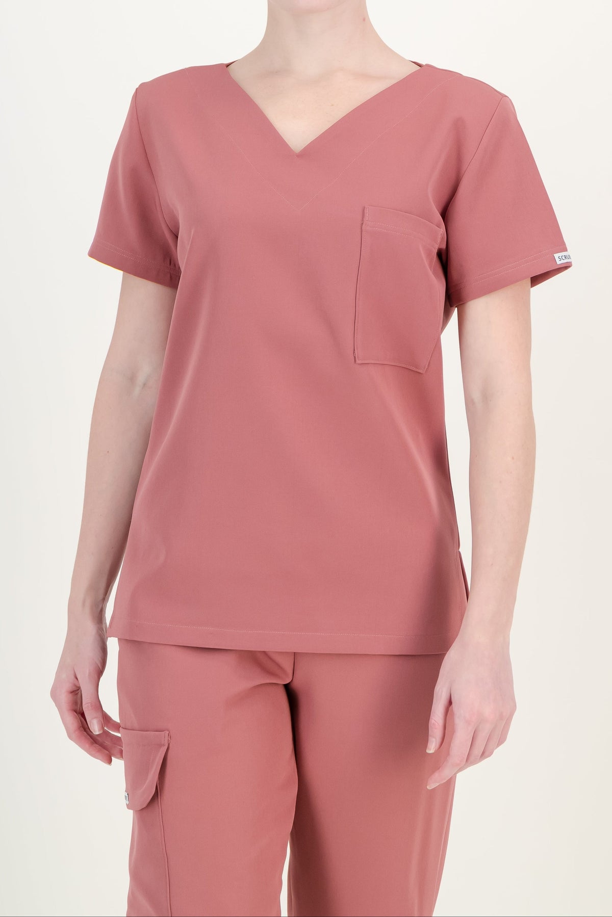 A woman modeling a dusty rose V-neck scrub top and matching scrub pants against a plain off-white studio background.